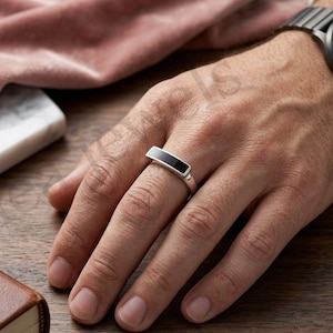 Puede incluir: Un anillo de tono plateado con una incrustación plana y rectangular de material negro liso en la parte superior. La banda tiene un acabado metálico pulido y un diseño elegante de bordes rectos que se asienta horizontalmente.