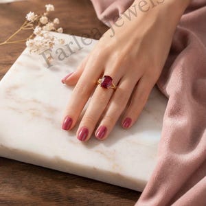 May include: A gold ring featuring a large, rectangular, deep red gemstone set in a prong setting. The ring is worn on a finger, showcasing the vibrant red stone against a polished gold band. The text Farlea jewels is visible in the image.