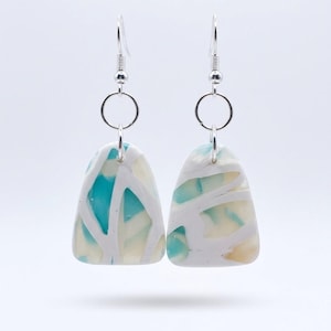 May include: A pair of dangle earrings featuring triangular-shaped pendants made of white, turquoise, and cream-colored marbled material. The pendants are suspended from silver-toned metal fishhook wires by small circular jump rings.