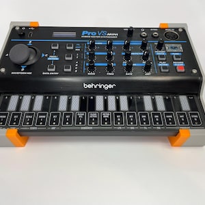 May include: A black Behringer Pro VS Mini hybrid vector synthesizer with a gray and orange stand. The device features a joystick for waveform mixing, multiple control knobs, a small display screen, and a touch-sensitive keyboard interface.