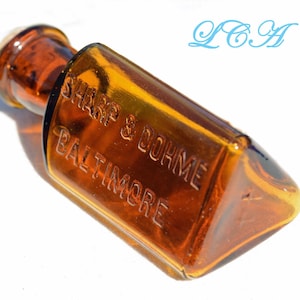 Puede incluir: Una botella de vidrio color ámbar con un tapón marrón. La botella rectangular tiene bordes redondeados y texto en relieve que dice "SHARP & DOHME BALTIMORE". La botella está acostada de lado.