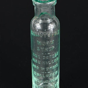 May include: A clear glass bottle with green lettering. The bottle reads "Rumsteads Worm Syrup" and other text. The bottle has a rounded body and a narrow neck. The bottle is empty and appears to be an antique.