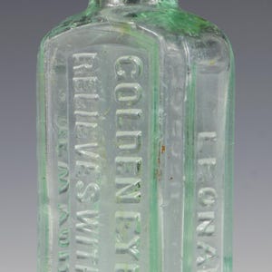 Puede incluir: Botella de vidrio transparente antigua con texto en relieve. La botella es rectangular con esquinas redondeadas y un cuello estrecho. El texto dice "Golden Eye Lotion" y "Relieves Without Pain". La botella tiene un ligero tinte verde.