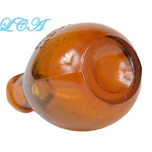 May include: Amber-colored glass bottle with a rounded body and a short, cylindrical neck. The bottle has a glossy finish and appears to be empty. The text "LCA" is visible in light blue on the upper left side of the bottle.