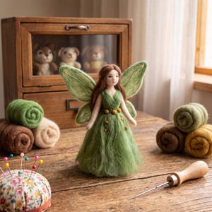May include: A needle-felted fairy figurine with long brown hair, large green wings, and a flowing green dress adorned with small yellow accents. The figure stands alongside rolls of wool, a floral pincushion, and a wooden felting tool.