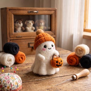 Puede incluir: Una figura de fantasma blanco de lana fieltrada con un gorro de punto naranja con pompón, que sostiene un pequeño cubo de calabaza naranja. El fantasma tiene ojos negros y mejillas rosadas. Cerca hay rollos de lana, un alfiletero y una aguja.