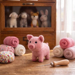 May include: A needle-felted pink pig figurine stands on a wooden surface next to a floral pincushion with colorful pins, a wooden-handled felting needle, and several rolls of pink and white wool roving, with felted animal figures in the background.