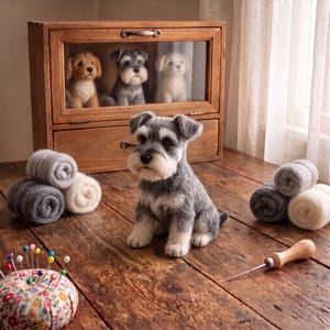 Pode incluir: Uma figura de cão Schnauzer em lã feltrada cinzenta e branca sobre uma superfície de madeira. Atrás, uma vitrine de madeira contém três figuras de cães mais pequenas. Há rolos de lã, uma almofada de alfinetes floral e uma ferramenta de feltragem.