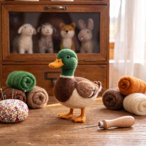 May include: A needle-felted mallard duck figurine with a green head, brown chest, and cream-colored body. Beside it are rolls of colored wool roving, a floral-patterned pincushion with colorful pins, and a wooden-handled felting needle.