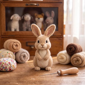 May include: A needle-felted beige rabbit figurine stands upright. Surrounding the rabbit are several rolls of wool roving in shades of cream, tan, and brown, a floral print pincushion with colourful pins, and a wooden-handled felting needle.