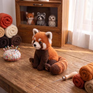May include: A needle-felted red panda figurine with orange and dark brown wool, featuring a bushy striped tail. Beside it sits a floral pincushion with colorful pins and a wooden-handled felting needle on a rustic wooden surface.