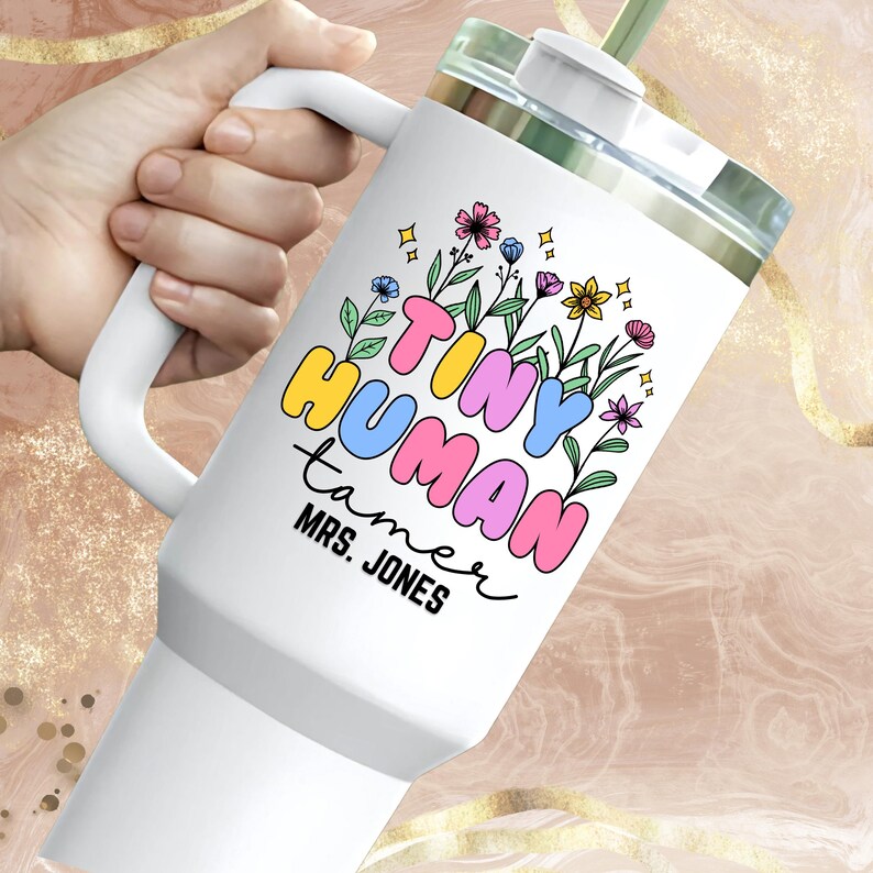 May include: White tumbler with a black and colorful design that says "Tiny Human Tamer" with the name "Mrs. Jones" underneath. The tumbler has a lid and a straw.