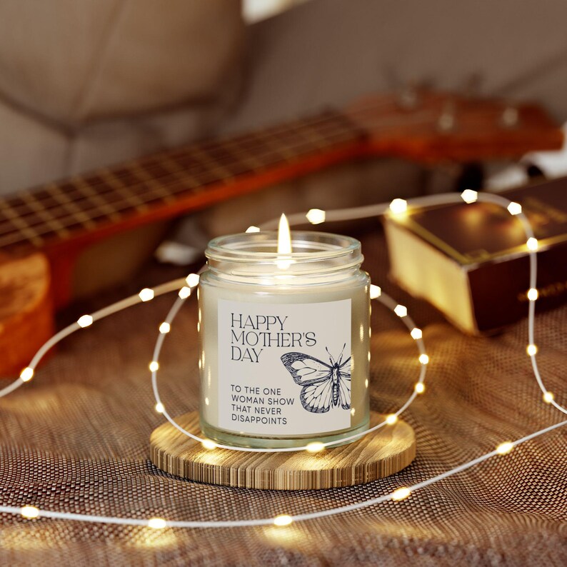 Peut inclure: Une bougie blanche dans un pot en verre avec une &eacute;tiquette qui dit "Happy Mother's Day to the one woman show that never disappoints". La bougie est allum&eacute;e et il y a un graphique de papillon sur l'&eacute;tiquette.