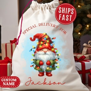 May include: A cream-colored Christmas gift bag with a drawstring closure. It features a gnome illustration wearing a red hat and a string of colorful lights. Text on the bag reads "Special Delivery For Jackson". A red circle reads "Ships Fast".