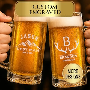 May include: Two clear glass beer mugs filled with golden lager, each featuring custom white engraving. The left mug displays the name JASON, a mountain graphic, and the text BEST MAN JULY 12, 2026. The right mug shows the letter B, antlers, and BRANDON GROOMSMAN.