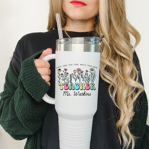 May include: White stainless steel travel mug with a black lid and a handle. The mug has a colorful graphic of flowers and the text "Teach them, love them, watch them grow. Teacher Ms. Watkins".