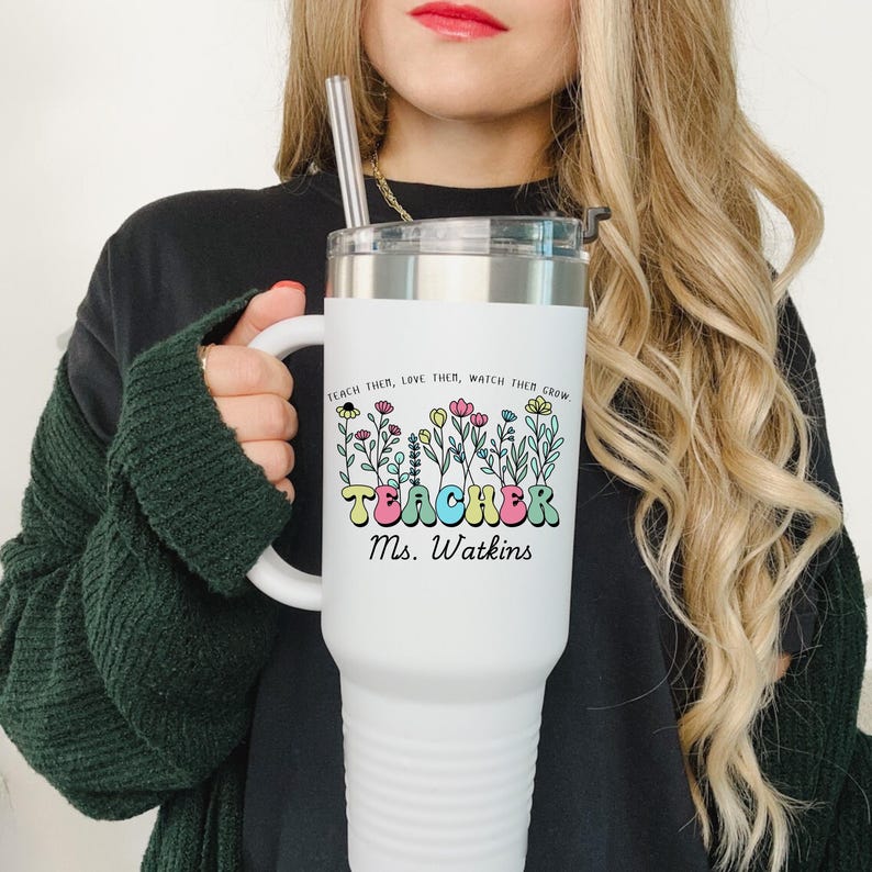 May include: White stainless steel travel mug with a black lid and a handle. The mug has a colorful graphic of flowers and the text "Teach them, love them, watch them grow. Teacher Ms. Watkins".