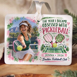 May include: A rectangular white ornament with a white ribbon hanger featuring a photo of a person playing pickleball on the left and text on the right that reads THE YEAR I BECAME OBSESSED WITH PICKLEBALL, JENNA 2026, and BOULDER PICKLEBALL CLUB.