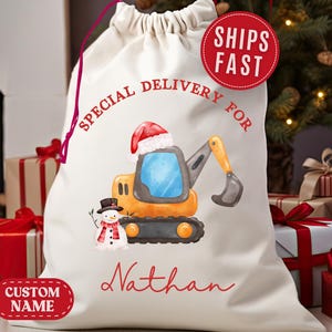 May include: A cream-colored Christmas gift bag with a drawstring closure. The bag features a cartoon excavator wearing a Santa hat and a snowman. Text on the bag reads "Special Delivery For" and "Nathan". A red circle reads "Ships Fast".