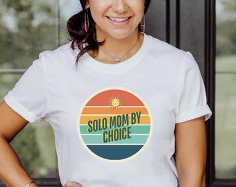 May include: White t-shirt with a retro-style graphic of a sun and the text "Solo Mom By Choice".