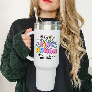 May include: White stainless steel tumbler with a black handle and a colorful graphic that says "Tiny Human Tamer" with the name "Mrs. Jones" underneath. The tumbler has a straw and is decorated with flowers.