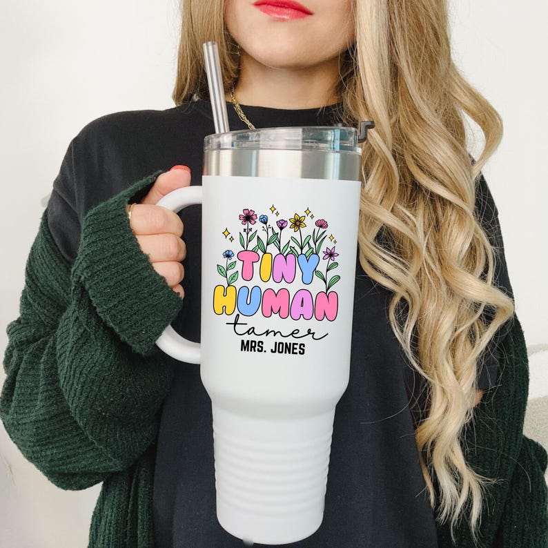 May include: White stainless steel tumbler with a black handle and a colorful graphic that says "Tiny Human Tamer" with the name "Mrs. Jones" underneath. The tumbler has a straw and is decorated with flowers.