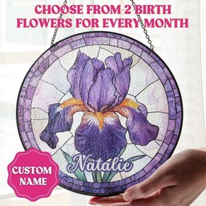 May include: Round stained glass window art featuring a purple iris flower with yellow accents. The design includes the text "Choose from 2 birth flowers for every month" and a custom name. The window is held by a hand.