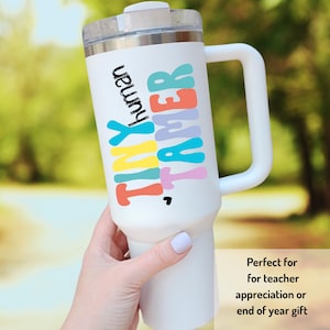 May include: White travel mug with a black handle. The mug has colorful text that reads "Tiny Tamer of human".