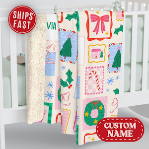 May include: A Christmas-themed blanket with a cream-colored background and a repeating pattern of festive elements. The design includes Christmas trees, bows, candy canes, and houses in pink, green, and blue. The blanket has a soft, cream-colored sherpa lining.