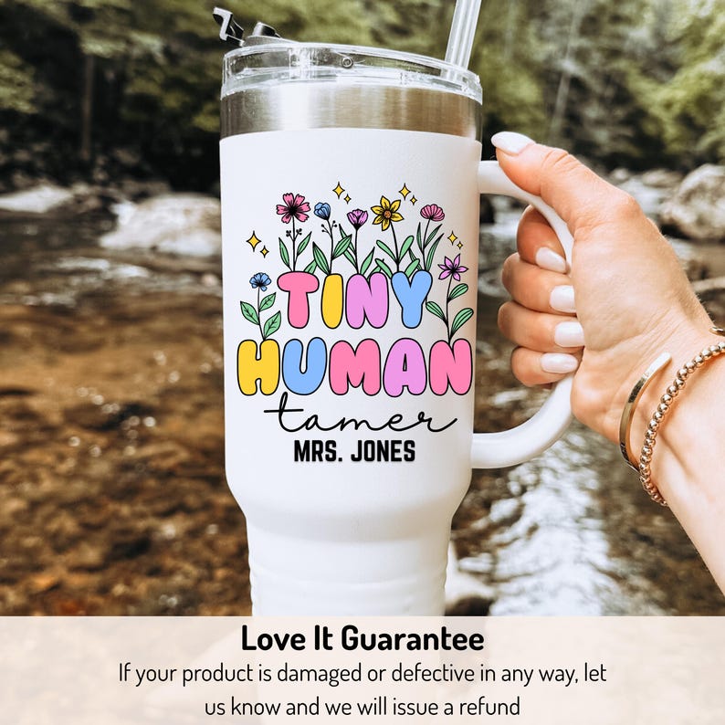 May include: White stainless steel travel mug with a colorful graphic that says "Tiny Human Tamer" and "Mrs. Jones". The graphic features flowers and stars.
