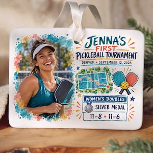 May include: A rectangular white ceramic ornament with a white ribbon hanger. It features a photo of a smiling person holding a pickleball paddle, alongside text reading Jenna's First Pickleball Tournament, Denver, September 12, 2026, Women's Doubles, Silver Medal, and the scores 11-8, 11-6.