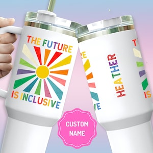 May include: Two white stainless steel tumblers with lids and straws. The tumbler on the left has a rainbow sunburst design and the text "The Future is Inclusive". The tumbler on the right has the text "Heather" and a rainbow sunburst design. The tumblers are on a pink and blue background.