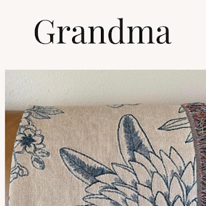 Puede incluir: Una manta de tejido personalizada con un dise&ntilde;o floral y la palabra "Abuela" en cursiva. La manta es beige con detalles florales azules y tiene flecos en los bordes.