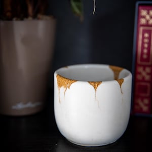 May include: A white ceramic cup with a gold-colored rim and drips. The cup is round and sits on a black surface. A brown plant pot and a red book are in the background.
