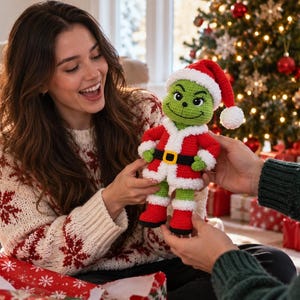 Op de afbeelding: Een gehaakte pop van een groen personage met een puntig gezicht, grote ogen en een brede glimlach. De pop draagt een rood kerstmanpak met witte randen, een zwarte riem met een gele gesp en rode laarzen met witte boorden, inclusief een rode kerstmuts.
