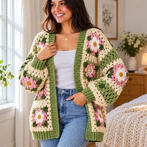 Puede incluir: Un cárdigan de ganchillo de manga larga con un patrón de cuadrados de la abuela con motivos florales en crema, verde salvia y rosa. La prenda tiene un borde acanalado de color verde salvia en la abertura frontal, puños y el dobladillo.