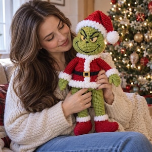 May include: A crocheted green character doll wearing a red and white Santa suit with a matching hat. The doll features a stitched facial expression with arched eyebrows, a black belt with a gold buckle, and red boots with white trim cuffs.