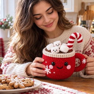Puede incluir: Una taza roja de ganchillo con una cara sonriente, mejillas rosadas y detalles de copos de nieve blancos. La taza está rellena de líquido marrón de ganchillo, tres malvaviscos blancos sonrientes y un bastón de caramelo a rayas rojas y blancas.
