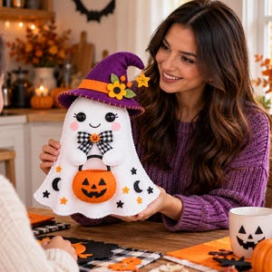 May include: A white felt ghost decoration wearing a purple witch hat with orange trim and yellow flower accents. The ghost features black eyes, a small smile, a black and white checkered bow, and holds an orange felt jack-o'-lantern bucket.