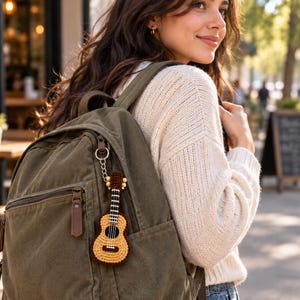 May include: An olive green canvas backpack featuring a small, crocheted guitar keychain attached to the zip. The keychain is crafted with yellow and brown yarn, depicting a guitar with a dark brown neck, white fret markers, and a round body.