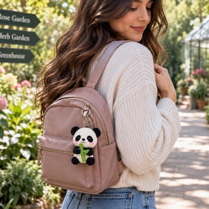 May include: A dusty rose mini backpack featuring a gold-tone zipper and a small, crocheted panda bear keychain attached to the front. The panda is white and black with pink cheeks, holding a small green bamboo stalk in its arms.