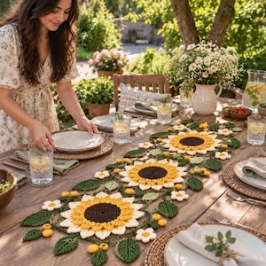 May include: A crochet table runner featuring large sunflowers with dark brown centers, yellow petals, and green leaves, accented by smaller white and yellow flowers, laid out on a wooden dining table set with plates, glasses, and bowls.