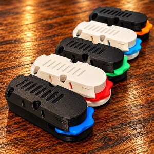 May include: A series of five oval-shaped plastic cable organizers arranged in a diagonal line. Each unit features a top cover with parallel ventilation slots and a bottom base with a scalloped edge in blue, red, green, white, or orange.