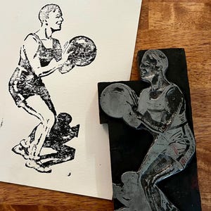 Peut inclure: Une impression en noir et blanc d'une personne jouant au basket-ball, tenant un ballon. L'impression est sur papier blanc. &Agrave; c&ocirc;t&eacute;, un bloc noir avec la m&ecirc;me image, probablement un bloc d'impression.