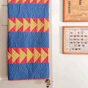 Könnte beinhalten: Ein rechteckiger Quilt mit abwechselnden horizontalen Streifen aus einfarbig blauem Stoff und Reihen aus gelben und roten Dreiecksmustern. Der Quilt hat eine texturierte, gesteppte Oberfläche und hängt vertikal an einer Tür.
