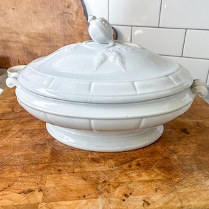 May include: A white ceramic soup tureen with a lid featuring a bird-shaped handle and leaf detailing. The tureen has a pedestal base and is set on a wooden cutting board. The handles are curved and the tureen has a basket weave design.