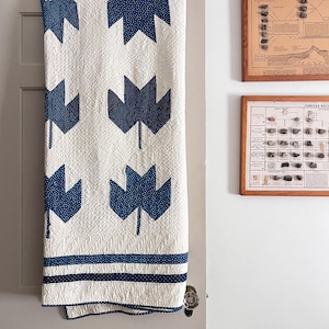Peut inclure: Une courtepointe blanche ornée d'un motif répétitif de feuilles d'érable bleu foncé avec un imprimé à petits points blancs. La courtepointe présente une surface matelassée texturée et deux bordures horizontales à rayures bleu foncé.