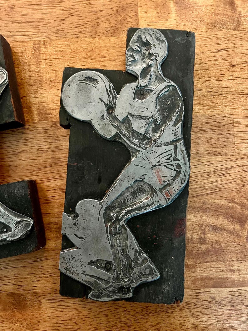 Peut inclure: Bloc d'impression en m&eacute;tal vintage repr&eacute;sentant un joueur de basket-ball en action. La figurine argent&eacute;e est mont&eacute;e sur un bloc de bois noir rectangulaire. Le joueur tient un ballon de basket. Le bois est vieilli.
