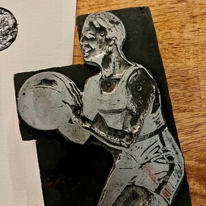 Peut inclure: Sculpture en relief en m&eacute;tal argent&eacute; repr&eacute;sentant un joueur de basket-ball en action, sur fond noir. Le joueur porte un d&eacute;bardeur et un short, tenant un ballon de basket. La sculpture est mont&eacute;e sur une base rectangulaire.