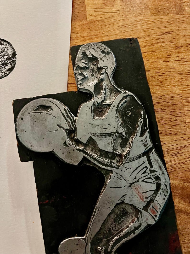 Peut inclure: Sculpture en relief en m&eacute;tal argent&eacute; repr&eacute;sentant un joueur de basket-ball en action, sur fond noir. Le joueur porte un d&eacute;bardeur et un short, tenant un ballon de basket. La sculpture est mont&eacute;e sur une base rectangulaire.